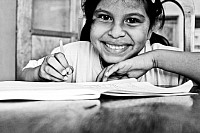 A young girl smiling while seated at a desk. She is leaning over an opened school book on the desk