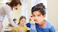 A child seated at a desk looking straight into the camera with a hopeless look on her face